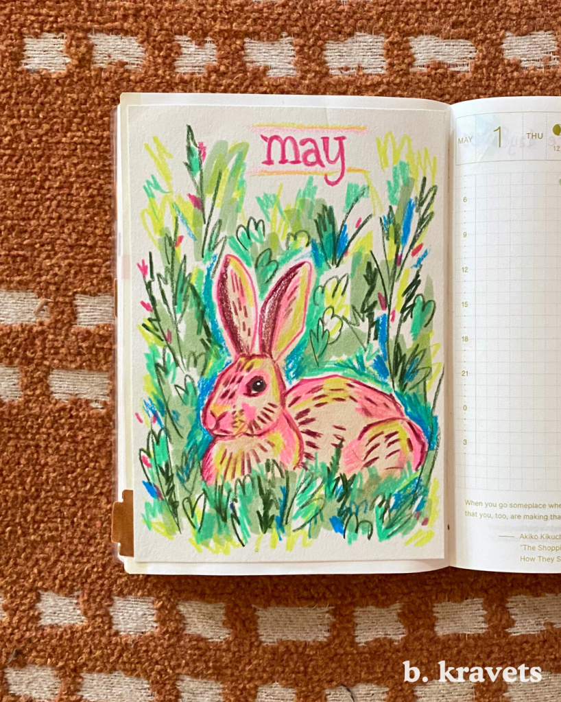 A hobonichi planner lays open on a blanket. On the left page is monthly title page with a marker and colored pencil drawing of a rabbit laying in the grass with the title “may” at the top.  The drawing is done in a loose scribbly style with vibrant and neon colors, mostly pinks and greens.