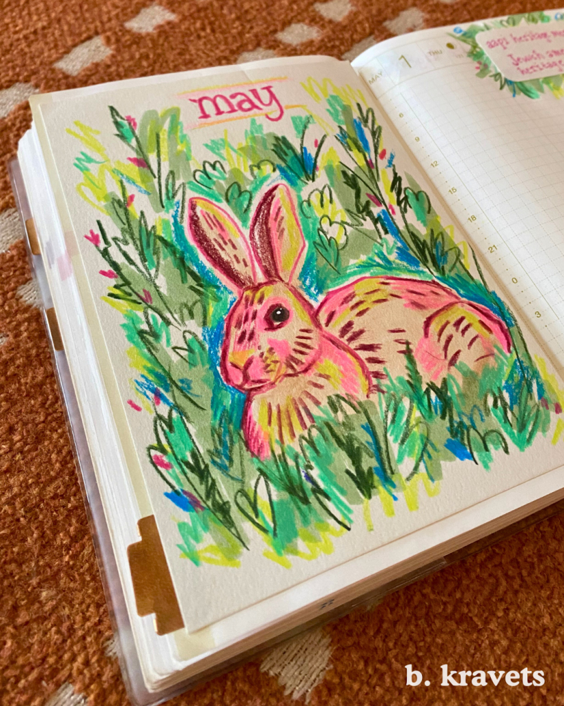 A hobonichi planner lays open on a blanket. On the left page is monthly title page with a marker and colored pencil drawing of a rabbit laying in the grass with the title “may” at the top.  The drawing is done in a loose scribbly style with vibrant and neon colors, mostly pinks and greens.