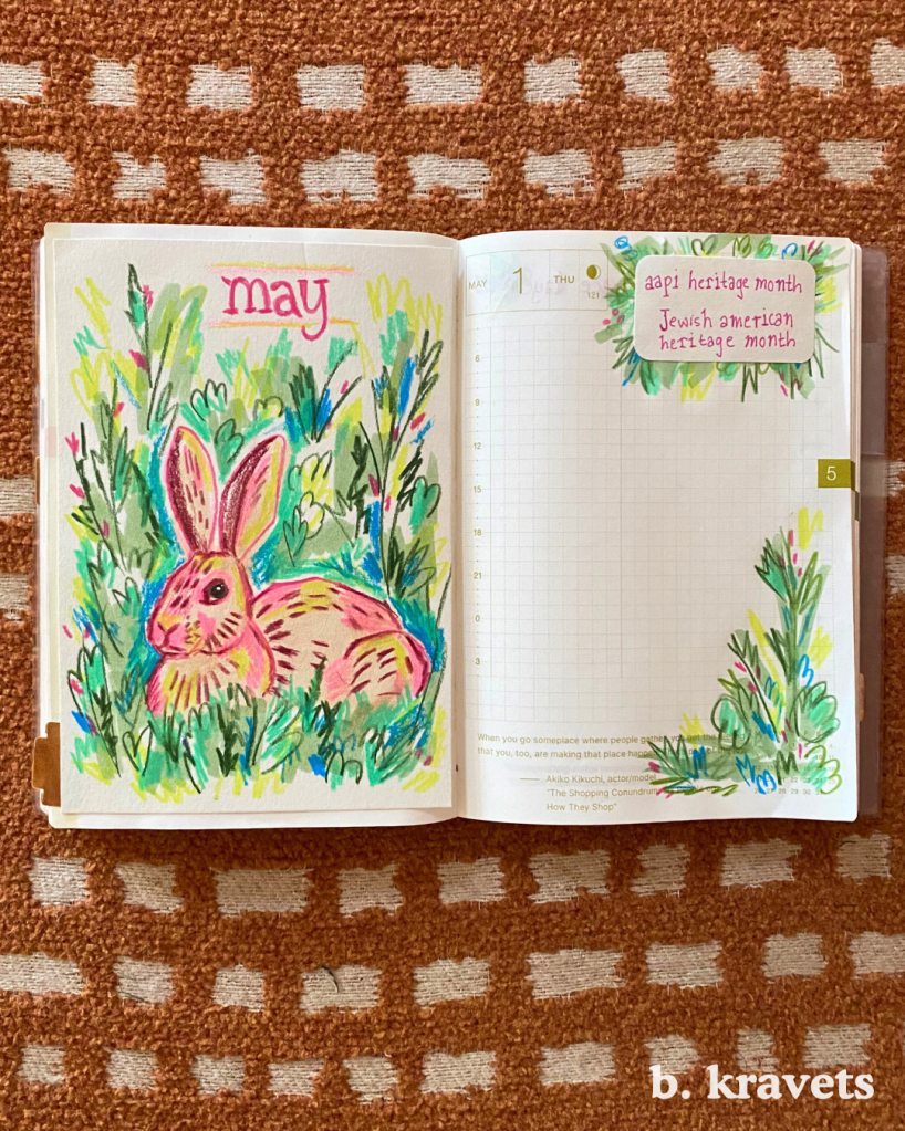 A hobonichi planner lays open on a blanket. On the left page is monthly title page with a marker and colored pencil drawing of a rabbit laying in the grass with the title “may” at the top.  The drawing is done in a loose scribbly style with vibrant and neon colors, mostly pinks and greens. The right page is the daily page for May 1 and has the titles “AAPI Heritage month” and “Jewish American Heritage month” at the top with drawings of foliage in the same style as the left page.