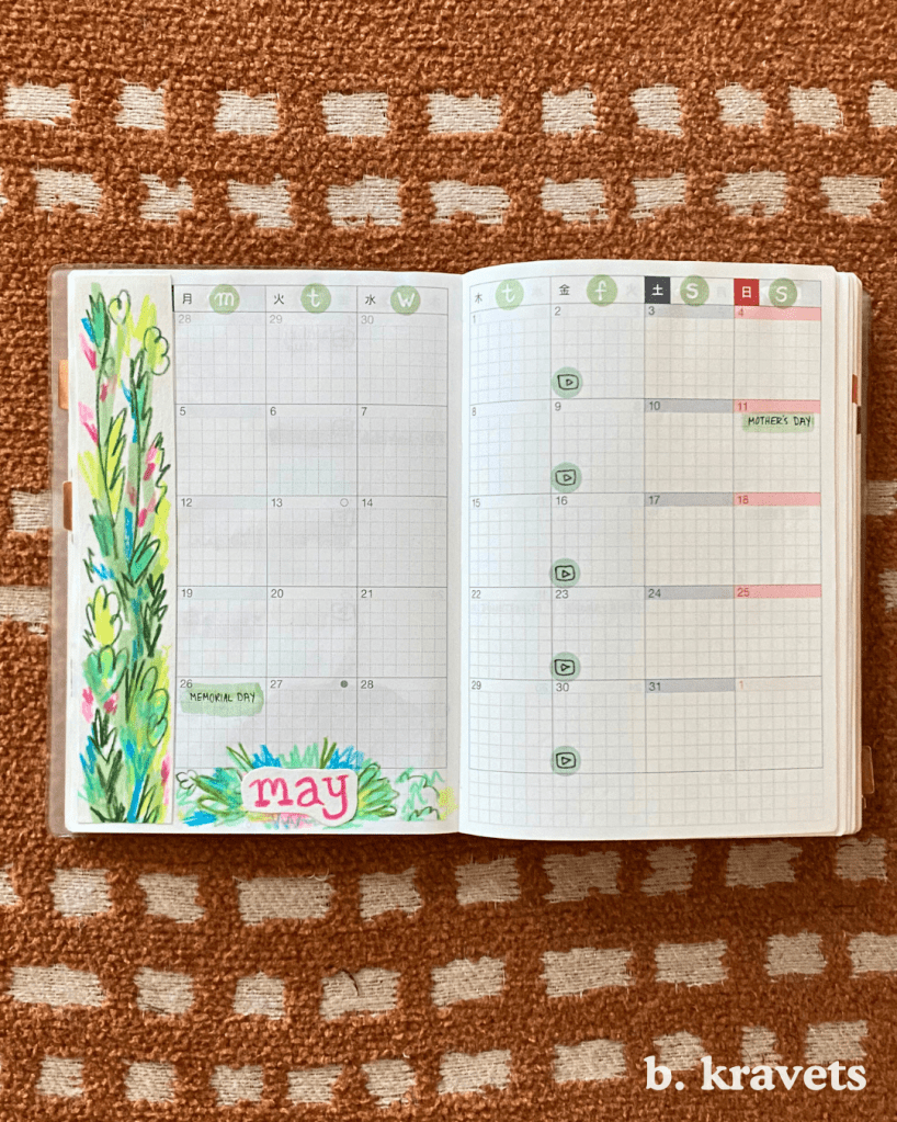 A hobonichi planner lays open on a blanket. It is open tot the May monthly calendar. The left side of the calendar is decorated with a drawing of foliage with the title “may” at the bottom. The drawing is done in a loose scribbly style with vibrant and neon colors, mostly pinks and greens.